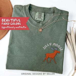 May include: An olive green t-shirt with the embroidered phrase "SILLY MOOSE" and a cartoon moose graphic. The shirt also features the text "BEAUTIFUL FADED COLORS Effortlessly aesthetic" and "ORIGINAL DESIGNS BY SELLER". A gold necklace with a pendant is draped on the shirt.