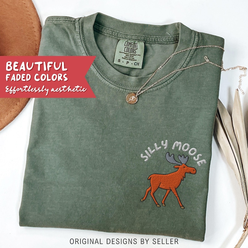 May include: An olive green t-shirt with the embroidered phrase "SILLY MOOSE" and a cartoon moose graphic. The shirt also features the text "BEAUTIFUL FADED COLORS Effortlessly aesthetic" and "ORIGINAL DESIGNS BY SELLER". A gold necklace with a pendant is draped on the shirt.