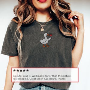 May include: Dark gray t-shirt featuring an embroidered cartoon duck holding a glass of red wine. The duck is gray with orange feet and beak. The shirt has a vintage, washed look.