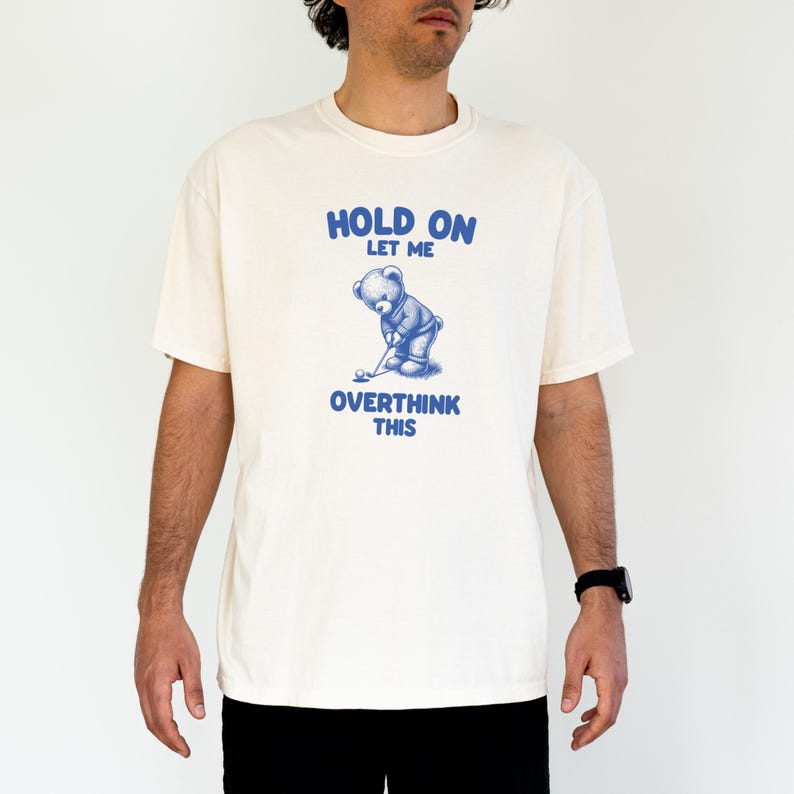 K&ouml;nnte beinhalten: Ein wei&szlig;es T-Shirt mit einer blauen Grafik eines Teddyb&auml;ren, der Golf spielt. Der Text "HOLD ON LET ME OVERTHINK THIS" ist auf das Shirt gedruckt.