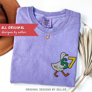 May include: A lilac Comfort Colors t-shirt with a cartoon goose holding the number 67. The shirt has a crew neck and short sleeves. A gold necklace with a pendant is draped on the shirt. The text "ALL ORIGINAL designs by seller" is visible.