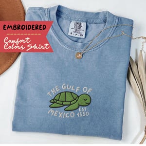 May include: A light blue t-shirt with an embroidered design of a sea turtle with the text "The Gulf of Mexico Est 1550".