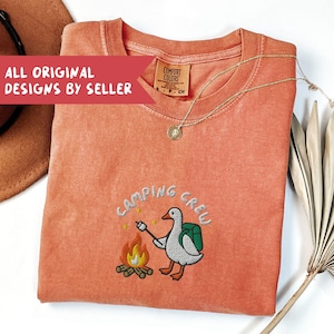 Op de afbeelding: Koraalrood T-shirt met de geborduurde tekst "CAMPING CREW" en een cartoon gans die een marshmallow roostert boven een kampvuur. Het shirt heeft een ronde hals en een klein label. Een gouden ketting en een hoed zijn ook zichtbaar.