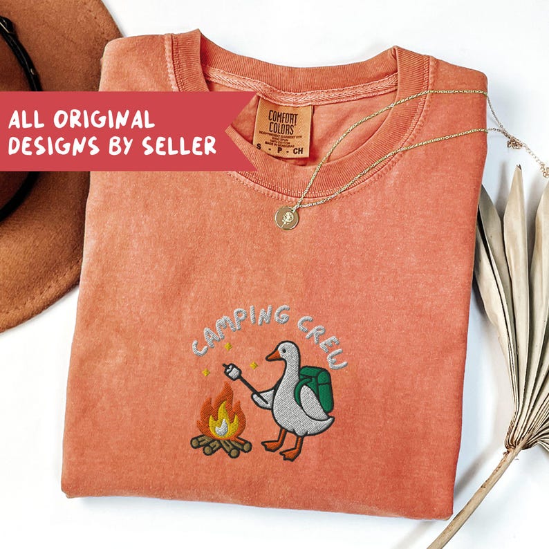 Op de afbeelding: Koraalrood T-shirt met de geborduurde tekst "CAMPING CREW" en een cartoon gans die een marshmallow roostert boven een kampvuur. Het shirt heeft een ronde hals en een klein label. Een gouden ketting en een hoed zijn ook zichtbaar.