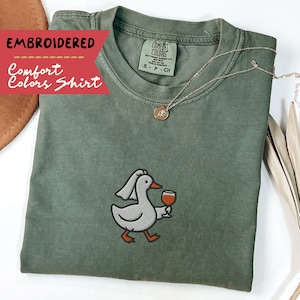 May include: A sage green Comfort Colors t-shirt with an embroidered duck design, wearing a veil and holding a wine glass. The shirt has a crew neck and short sleeves. A gold necklace with a pendant is also visible.