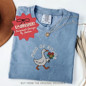 May include: A light blue t-shirt with the embroidered text "MAID OF HONOR" and a cartoon goose holding flowers. The shirt has a Comfort Colors label and a gold necklace. A red tag reads "EMBROIDERY Original Design by Seller".