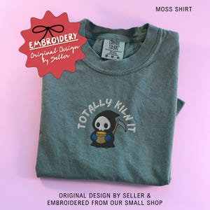 May include: A moss green t-shirt featuring an embroidered graphic of a cartoon grim reaper holding a yellow pot with the text "TOTALLY KILN IT" arched above. The shirt has a crew neckline and a Comfort Colors brand label inside the collar.