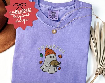 May include: A light purple Comfort Colors t-shirt with an embroidered design. The design features a ghost wearing an orange beanie and holding a turkey, with the word "THANKFUL" above it. A gold necklace is draped across the shirt.