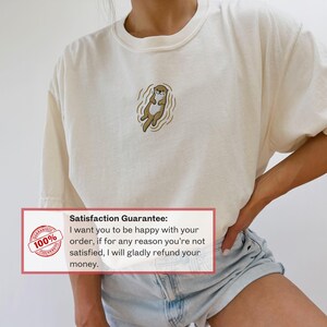 May include: Cream-colored t-shirt featuring an embroidered otter design. The otter is depicted in shades of brown and tan, floating in water. The shirt has a relaxed fit. The image also includes a guarantee.