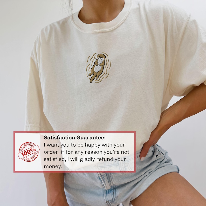 May include: Cream-colored t-shirt featuring an embroidered otter design. The otter is depicted in shades of brown and tan, floating in water. The shirt has a relaxed fit. The image also includes a guarantee.