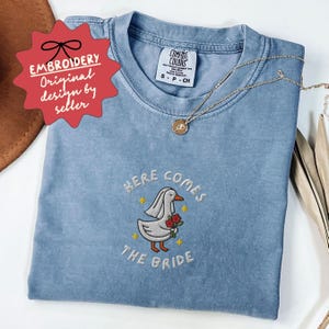 May include: A light blue t-shirt with the embroidered phrase "HERE COMES THE BRIDE" and a cartoon duck wearing a veil and holding flowers. The shirt also has a small tag that says "COMFORT COLORS". A gold necklace is draped across the shirt.