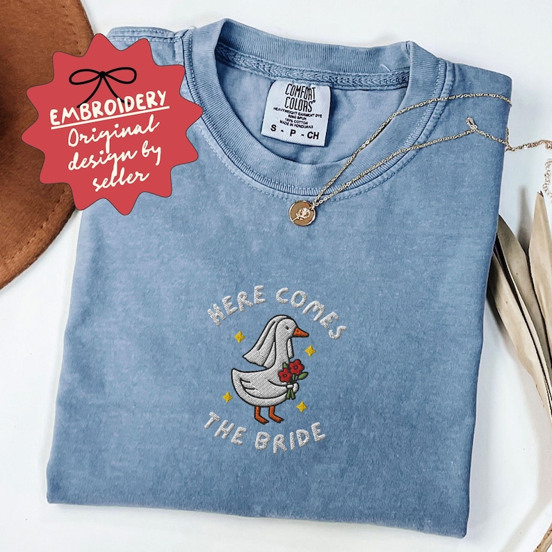 May include: A light blue t-shirt with the embroidered phrase "HERE COMES THE BRIDE" and a cartoon duck wearing a veil and holding flowers. The shirt also has a small tag that says "COMFORT COLORS". A gold necklace is draped across the shirt.