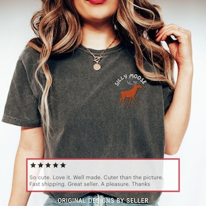 May include: Dark gray t-shirt with a small embroidered design of a moose and the words "Silly Moose". The shirt has a crew neck and short sleeves. The text "ORIGINAL DESIGNS BY SELLER" is at the bottom.