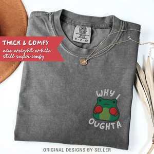 May include: A gray Comfort Colors t-shirt with an embroidered frog graphic and the text "WHY I OUGHTA." The shirt has a crew neck and short sleeves. A gold necklace with a pendant is draped on the shirt.