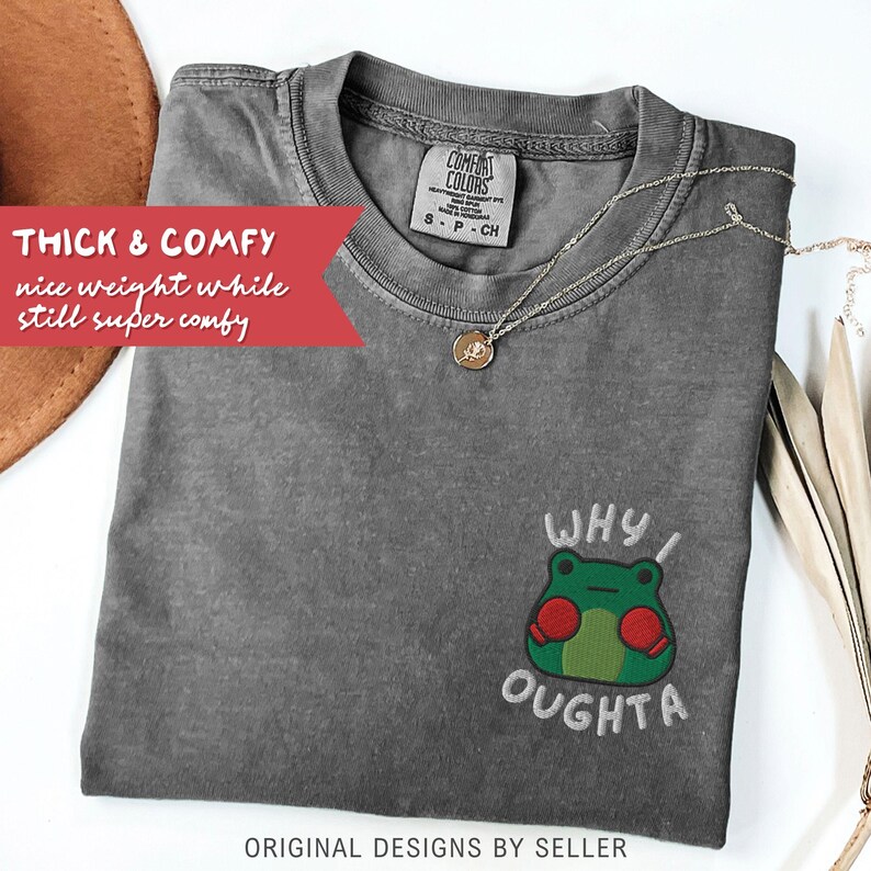 May include: A gray Comfort Colors t-shirt with an embroidered frog graphic and the text "WHY I OUGHTA." The shirt has a crew neck and short sleeves. A gold necklace with a pendant is draped on the shirt.
