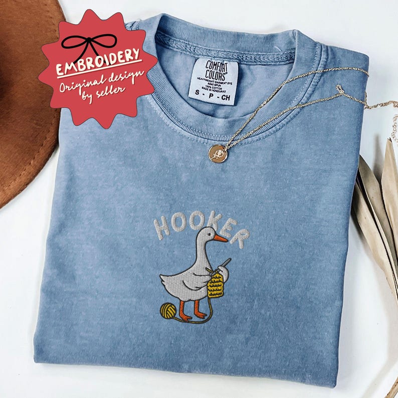 May include: A light blue t-shirt with the word "HOOKER" embroidered above a cartoon goose knitting. The shirt has a Comfort Colors tag and a gold necklace. A red tag reads "EMBROIDERY Original design by seller."