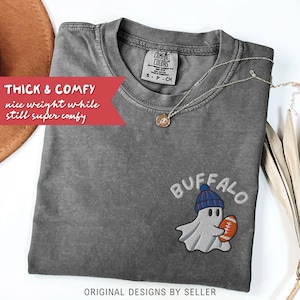 May include: A gray Comfort Colors t-shirt with an embroidered ghost wearing a blue and red hat and holding a football. The word "BUFFALO" is embroidered above the ghost. The text "THICK & COMFY" is on a red banner.