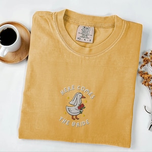 May include: Mustard yellow t-shirt with embroidered design. The design features a cartoon goose wearing a veil and holding red roses, with the text "HERE COMES THE BRIDE". A tag is visible on the collar.