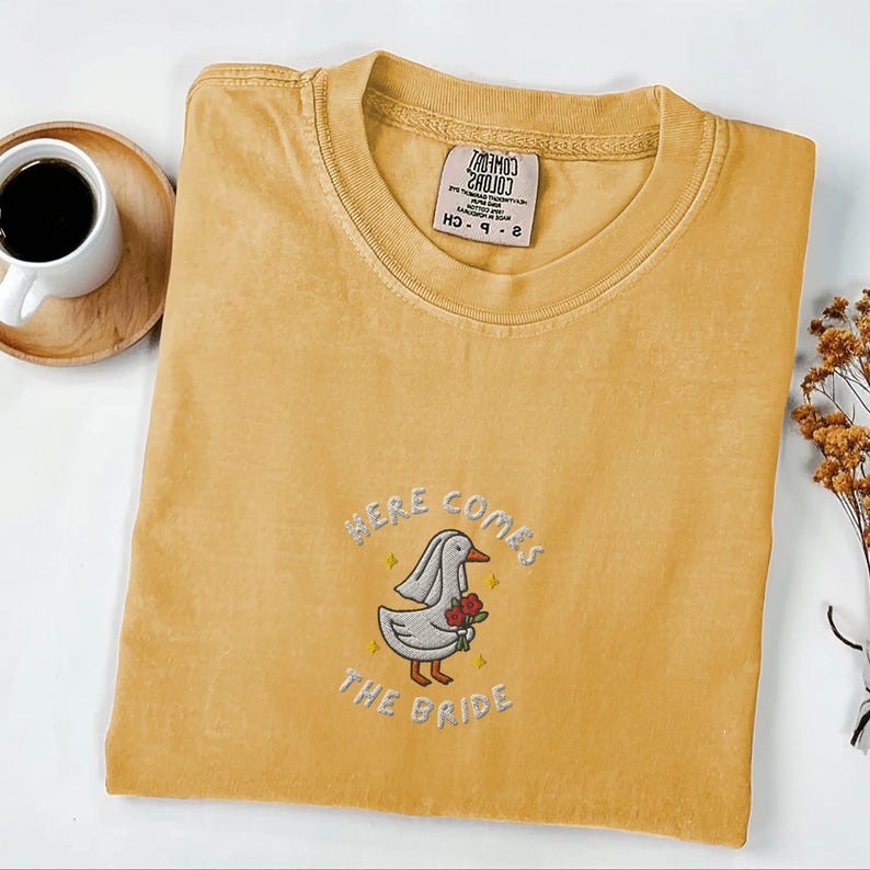 May include: Mustard yellow t-shirt with embroidered design. The design features a cartoon goose wearing a veil and holding red roses, with the text "HERE COMES THE BRIDE". A tag is visible on the collar.