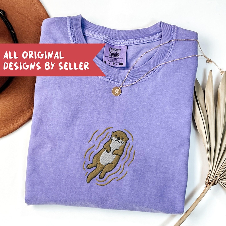 May include: A lavender Comfort Colors t-shirt featuring an embroidered otter design. The otter is brown and white, floating in water. A gold necklace with a pendant rests on the shirt. Text on a red banner reads "ALL ORIGINAL DESIGNS BY SELLER".