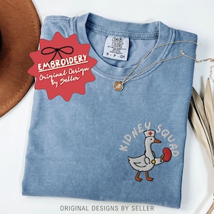 May include: A blue Comfort Colors t-shirt with an embroidered design featuring a cartoon goose wearing a nurse's hat and holding a kidney. The text "Kidney Squad" is embroidered above the goose. The shirt has a small tag that says "S-P-CH".