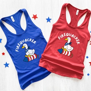 May include: Two racerback tank tops, one blue and one red, with a patriotic duck graphic. The duck wears a bandana and sits on a rocket with the word "FIREQUACKER" above. Red and blue star decorations are scattered around.