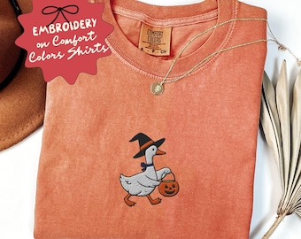 May include: An orange Comfort Colors shirt with an embroidered Halloween goose wearing a witch hat and carrying a pumpkin bucket. The shirt has the text "EMBROIDERY on Comfort Colors Shirts" on a red tag.