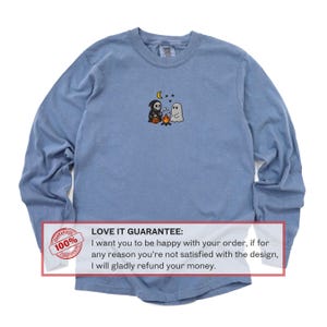 May include: A long-sleeve, light blue shirt with a small embroidered design of a grim reaper, a ghost, a campfire, a moon, and stars. The shirt has a crew neck and a tag on the back. The text "LOVE IT GUARANTEE" is also visible.