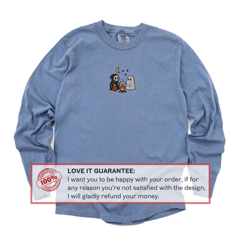 May include: A long-sleeve, light blue shirt with a small embroidered design of a grim reaper, a ghost, a campfire, a moon, and stars. The shirt has a crew neck and a tag on the back. The text "LOVE IT GUARANTEE" is also visible.