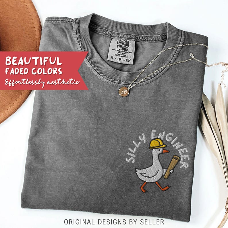 May include: A gray Comfort Colors t-shirt with the embroidered phrase "SILLY ENGINEER" and a cartoon goose wearing a yellow hard hat and carrying a blueprint. The shirt also has the text "ORIGINAL DESIGNS BY SELLER". A gold necklace is draped across the shirt.