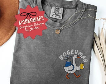 Peut inclure: Un t-shirt gris Comfort Colors avec un motif brodé. Le motif représente une oie de dessin animé portant des clubs de golf et une bière, avec le mot "BOGEYMAN" au-dessus. Le t-shirt porte également la mention "EMBROIDERY Original Design by Seller".