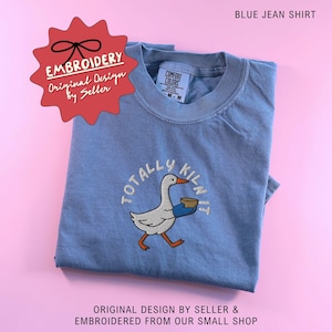 May include: A blue jean shirt with an embroidered design. The design features a cartoon goose holding a bowl, with the words "TOTALLY KILN IT" embroidered around it. The shirt also has the text "EMBROIDERY Original Design by Seller".