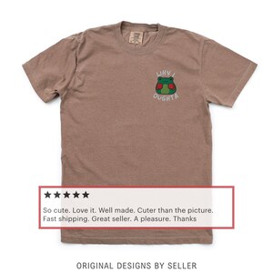 May include: Brown t-shirt with a small embroidered frog graphic. The frog has green skin, red cheeks, and the text "WHY I OUGHTA" above it. The shirt has a crew neck and short sleeves.