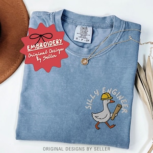 May include: A light blue Comfort Colors t-shirt with an embroidered design of a cartoon goose wearing a yellow hard hat and carrying blueprints. The text "SILLY ENGINEER" is embroidered above the goose. A gold necklace is draped on the shirt.