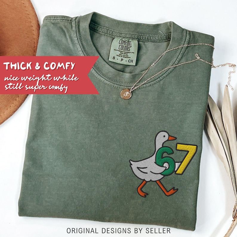 May include: Olive green Comfort Colors t-shirt with a cartoon goose graphic holding the number 67. The shirt has the text "THICK & COMFY" and "ORIGINAL DESIGNS BY SELLER". A gold necklace with a pendant is draped on the shirt.