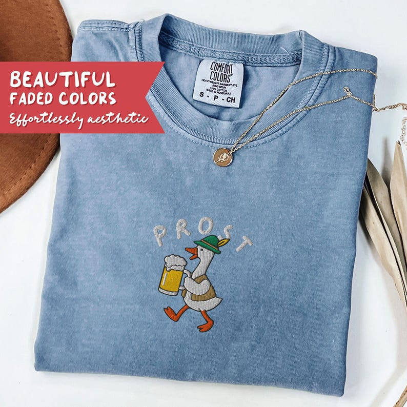 May include: A light blue Comfort Colors t-shirt with the word "PROST" embroidered above a cartoon goose holding a beer stein. The shirt has a crew neck and short sleeves. A gold necklace with a pendant is draped on the shirt.