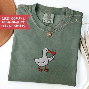 May include: Olive green Comfort Colors t-shirt featuring an embroidered cartoon duck holding a glass of red wine. A gold necklace with a pendant rests on the shirt. The text "COZY, COMFY & HIGH-QUALITY FEEL OF SHIRTS" is visible.