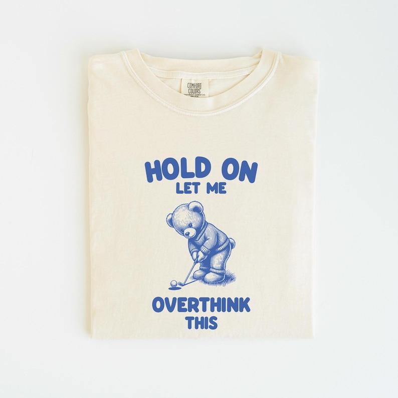 K&ouml;nnte beinhalten: Ein cremefarbenes T-Shirt mit einer blauen Grafik eines Teddyb&auml;ren, der Golf spielt. Der Text "HOLD ON LET ME OVERTHINK THIS" ist auf das Shirt gedruckt.