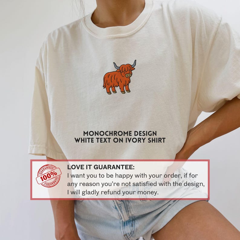 May include: Ivory-colored t-shirt featuring a cartoon illustration of a Highland cow in orange and brown. The shirt has the text "MONOCHROME DESIGN WHITE TEXT ON IVORY SHIRT". A guarantee is also shown.