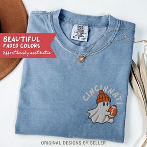 May include: A pale blue Comfort Colors t-shirt featuring the embroidered word "Cincinnati" above a cartoon ghost wearing an orange beanie and holding a football. The shirt has the text "Beautiful Faded Colors, Effortlessly aesthetic" and "Original Designs by Seller".