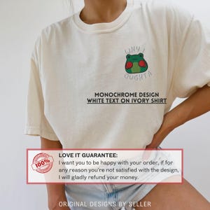 May include: Ivory-colored t-shirt featuring a green frog wearing red boxing gloves, with the text "WHY I OUGHTA." Below the graphic, the shirt reads "MONOCHROME DESIGN WHITE TEXT ON IVORY SHIRT." The shirt has a crew neck and short sleeves.