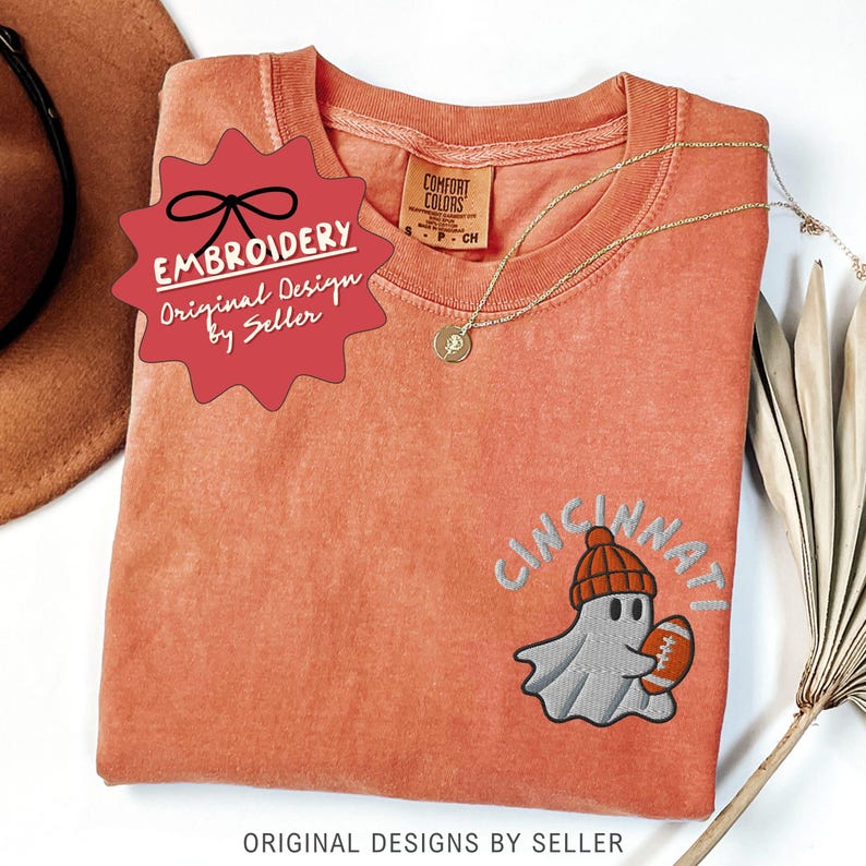 May include: Coral-coloured t-shirt with an embroidered design featuring a ghost wearing a beanie and holding a football, with the word "Cincinnati" above. A gold necklace and a red tag that says "Embroidery Original Design by Seller" are also visible.
