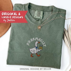 May include: Olive green t-shirt with an embroidered design of a goose wearing a lab coat and holding a medicine bottle, with the word "PHARMACIST" above it. The shirt also has the text "ORIGINAL & UNIQUE DESIGNS by Seller". A silver necklace is draped on the shirt.