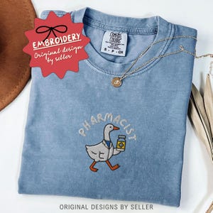 May include: A light blue Comfort Colors t-shirt with an embroidered design. The design features a cartoon goose wearing a lab coat and holding a medicine bottle, with the word "PHARMACIST" above it. A gold necklace is draped across the shirt.