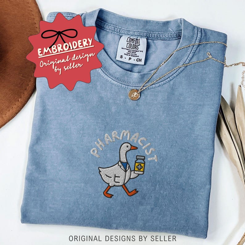 May include: A light blue Comfort Colors t-shirt with an embroidered design. The design features a cartoon goose wearing a lab coat and holding a medicine bottle, with the word "PHARMACIST" above it. A gold necklace is draped across the shirt.