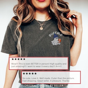 May include: Dark gray t-shirt with an embroidered design of a ghost wearing a blue beanie and holding a football. The word "BUFFALO" is arched above the ghost. Customer reviews are displayed below the design.