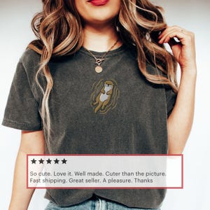 May include: Dark grey t-shirt featuring an embroidered otter design. The otter is depicted in a relaxed pose, with a light brown body and a cream-colored belly, surrounded by gold-colored wavy lines. The shirt has a classic crew neck.