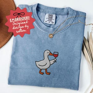 May include: A light blue Comfort Colors t-shirt featuring an embroidered cartoon duck holding a glass of red wine. The shirt has a crew neck and short sleeves. A gold necklace with a pendant is draped on the shirt. The tag reads "Embroidery Original design by seller."
