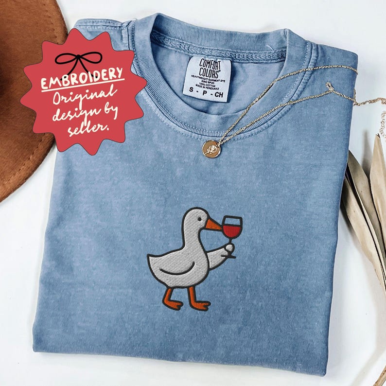 May include: A light blue Comfort Colors t-shirt featuring an embroidered cartoon duck holding a glass of red wine. The shirt has a crew neck and short sleeves. A gold necklace with a pendant is draped on the shirt. The tag reads "Embroidery Original design by seller."