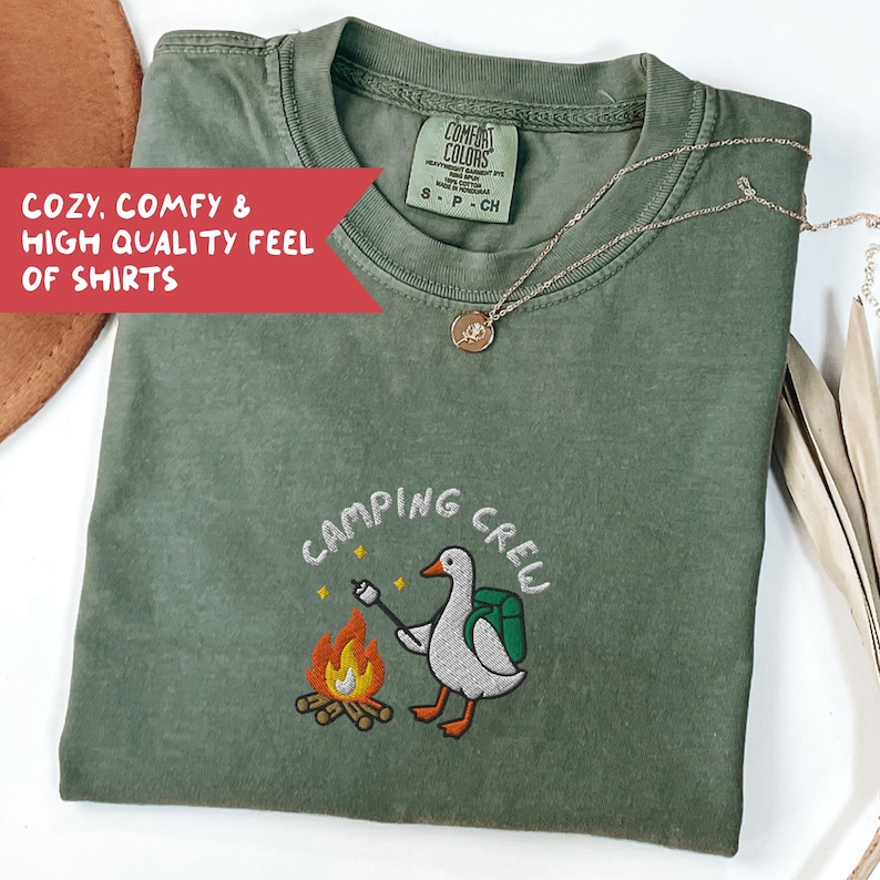 Op de afbeelding: Olijfgroen t-shirt met de geborduurde tekst "CAMPING CREW" en een cartoon gans die een marshmallow roostert boven een kampvuur. Het shirt heeft ook de tekst "COZY, COMFY & HIGH QUALITY FEEL OF SHIRTS". Een gouden ketting ligt over het shirt.
